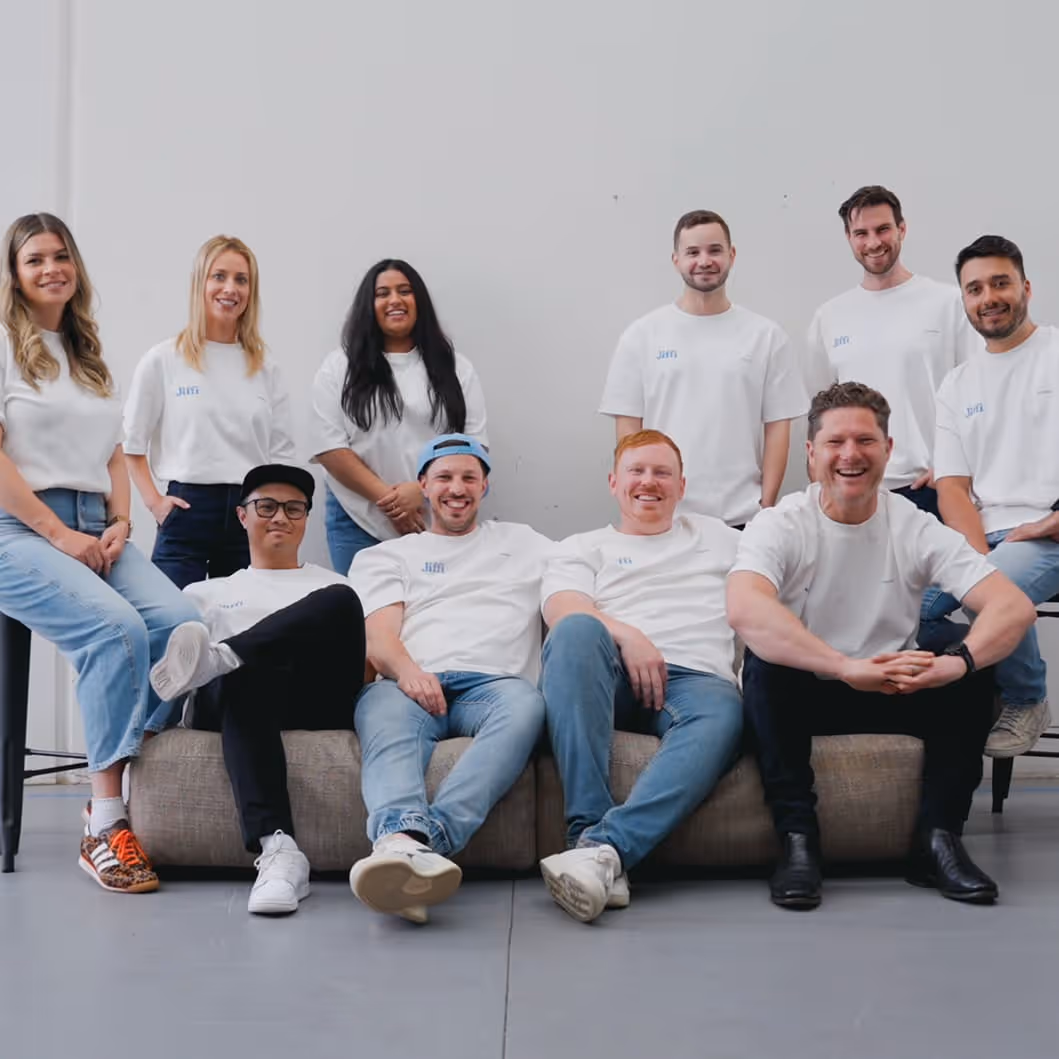 Group of ten people smiling and posing together indoors, wearing casual white shirts and jeans.